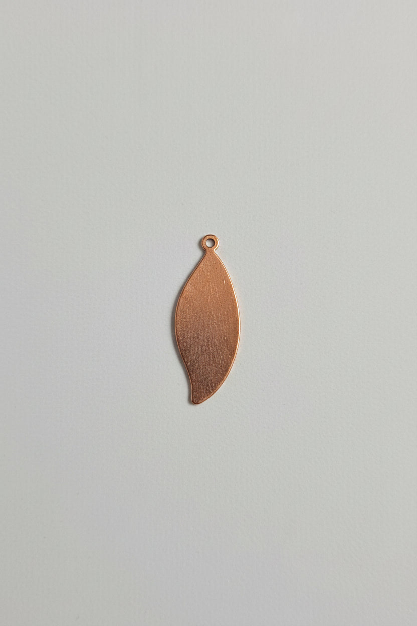A small copper-colored metal teardrop pendant with a loop at the top, shown on a black cutting mat with white grid lines and measurements.