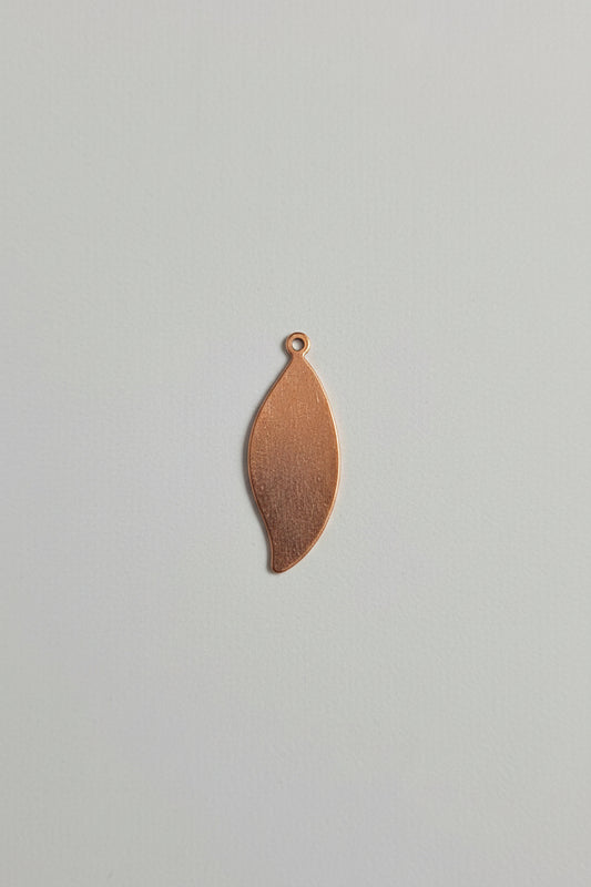 A small copper-colored metal teardrop pendant with a loop at the top, shown on a black cutting mat with white grid lines and measurements.